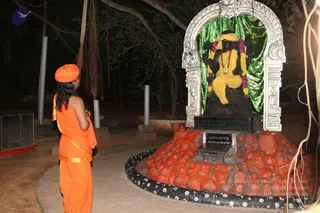 Night photograph at an outdoor shrine under dark trees. A figure in orange robes and an orange turban, seen from behind with long dark hair, stands with hands joined in prayer facing a black stone dakshinamurthy murti seated under a silver arched frame. The deity wears a yellow cloth, a rudraksha mala, and garlands of pink and purple flowers, and is set before a green backdrop. In front, a raised mound of red-orange kumkum or vermilion-coated stones surrounds the murti, with a small placed sign reading the deity's name and an inscription on the base. Hanging aerial roots frame the top of the scene, and metal poles stand in the background.