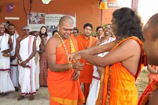 A photograph of an outdoor ceremony in front of an orange-washed building with a poster on the wall. A shaven-headed man in orange robes with a gold-bead necklace and gold bracelet smiles while another man with long dark hair, wearing orange robes and a gold-bead necklace, ties or adjusts a gold bracelet on his arm. Behind them stand several men in white dhotis with red-bordered shawls, a man in an orange kurta with glasses, and women in sarees, all watching. A woman in a pink saree stands at the left edge, and one more person is partially visible on the right. A poster beside them shows photographs and Kannada text.