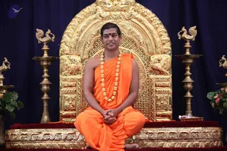 A seated man in saffron-orange robes sits cross-legged on an ornate golden throne with a carved arched backrest and lion-like carvings on the base. He wears a red bindi, a long beaded gold-toned necklace, and holds his hands clasped in his lap. Behind him hangs a deep blue draped backdrop, flanked by tall brass lamp stands topped with peacock-shaped finials, and a green plant with pink flowers sits at the right edge. The setting suggests a formal seated teaching or darshan.