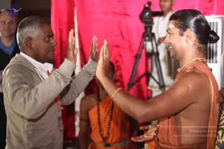 Two men face each other with palms pressed together in greeting. On the left, a grey-haired man in a beige zip hoodie and white collar stands with a lanyard and badge. On the right, a man with hair tied in a high bun, a white forehead mark and a gold necklace wears a draped saffron-orange shawl over bare shoulders and smiles. Behind them a seated person in orange robes with a long rudraksha mala sits in front of a red drape, and a camera on a tripod and a man in white clothing stand in the background. A white air conditioner unit is visible at right.