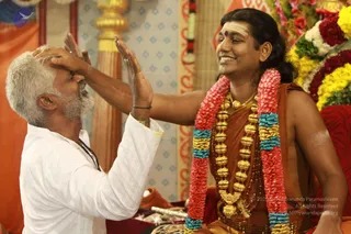 A photograph of a seated man in an orange-brown dhoti-style garment with a bare upper body, wearing a heavy gold necklace of large beads, a multicoloured garland of red, pink, yellow and teal flowers, and a red bindi with white sacred marks on his forehead. He smiles while a grey-haired, white-bearded man in a white shirt and dark prayer beads places both palms on the seated man's head in a blessing gesture. A red cushioned seat, floral decorations and a patterned backdrop with orange and gold panels surround them. Probable Swamiji cues: long swept-back hair, ornate gold necklace, saffron-orange clothing and rudraksha-style beads, receiving the blessing in a ceremonial setting, appears to be a seated blessing or darshan.
