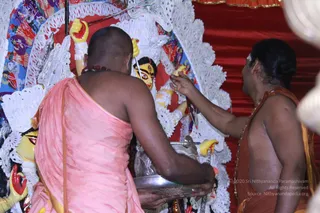 A photograph of a devotional rite at a large goddess murti with multiple raised yellow arms and a painted face, set against white lace-like decorative backdrop and a red cloth. Two men in ritual attire perform the worship: the man on the left is bald with a rudraksha mala, wearing a pink-salmon cloth wrapped around his body, and he holds a silver plate while reaching up toward the murti. The man on the right, bare-chested with a saffron-orange cloth and a beaded mala, touches the murti's arm with one hand. No distinctive a person cues are visible, so the people are described by role only.
