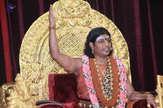A photograph of a man seated on an ornate gold throne with a carved golden backrest and a gold lion-shaped armrest, in front of deep red velvet drapes. He wears an orange-red garment, a rudraksha mala with large beads, a leopard-print shawl, a pink and white flower garland, gold earrings and a bangle, and a white sacred ash mark with a red bindi on his forehead. His right arm is raised in a gesture of address while he speaks. The Swamiji (probable) identity rests on his long swept-back hair, ornate gold seat, orange clothing, rudraksha mala and ceremonial gold ornaments, consistent with a discourse setting.