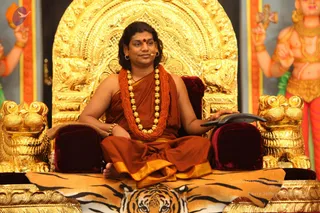 A man seated cross-legged on an ornate golden throne with lion-head armrests and a tiger-skin rug beneath him, speaking into a headset microphone. He wears an orange-red ochre robe with a gold border, a long orange-red beaded mala and a large gold bead necklace, with a red bindi on his forehead and gold earrings. He holds a small black device in one hand. Behind him, a golden arched backrest and painted deity figures with gold jewellery frame the scene. Swamiji (probable): long swept-back hair, saffron-ochre robe, rudraksha-style mala and gold necklace, seated on a throne as the speaker.