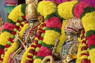 Close view of two golden metal deity murti decorated for a festival alankara, heavily adorned with yellow, pink and red flower garlands interspersed with green apples and reddish fruit. The figure on the left wears a tall golden crown and holds a golden staff wrapped in red and white cord; the figure on the right wears an ornate jewelled crown with red and white beadwork, a blue garment and a large green-and-red jewelled necklace. Ornate gold and gem jewellery covers both figures.