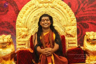 A smiling man with long, dark, swept-back hair sits on an ornate gilded throne with carved lion heads and a red-patterned backdrop. He wears a saffron-orange robe with a yellow wrap and multiple brown rudraksha malas, with a red tilak on his forehead, and his hands are clasped in his lap. Swamiji (probable): long swept-back hair, saffron robe and rudraksha malas on a throne, seated in person.
