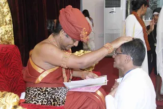 A photograph of a seated figure in a red-orange turban and matching orange-red robe with gold borders, wearing layered coral-coloured beaded necklaces and bracelets, placing a hand on the crown of a kneeling older man in white clothes and glasses. The seated figure holds an open book and a gold vessel on a red-cushioned throne with gold carved decoration behind. Two other people stand in white at the back, one wearing an orange shawl.