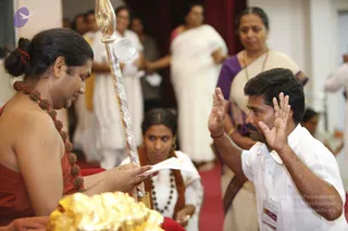 A photograph of a formal ceremony indoors with a red carpet. On the left, a man with hair tied in a bun wears an orange-red garment and a long rudraksha mala, and holds a small paper or card while seated beside a tall silver ceremonial staff topped with a round finial. In the foreground a gold-coloured ornate object is visible. On the right, a man in a white shirt with a lanyard raises both palms in a gesture of greeting or blessing. Behind them a woman in a purple-bordered sari and a woman in white saris stand, and another woman in a white outfit with a wooden-bead necklace kneels near the staff. Several other people in white are visible in the background. a person appears to be the figure in orange-red with the rudraksha mala, though the identification is uncertain.