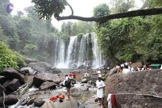 A jungle waterfall with several tiers of white water cascading down a dark rock face, surrounded by dense green trees and vines. In the foreground, a man in a white shirt and shorts stands on a rock with a dark backpack and a bag, and a few people in orange and white clothing wade in the shallow pool. Orange and red cloth lies on the rocks at the bottom left, and a small green-roofed shelter sits at the right edge. Several dozen people are scattered along the banks and in the water, many in white clothing, while a small group gathers on the right beside large boulders.