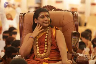 A man seated on an ornate brown leather throne with carved dark wooden arms and a cobra-head armrest carving on the right. He wears a saffron-orange robe with a gold-yellow edge, multiple strands of beads and a large gold ceremonial necklace, a red-and-blue wrist band, and a red tilak on his forehead. His long dark hair falls to his shoulders, and he rests one hand against his cheek while looking to the side. A small microphone is clipped near his face. Behind him, seated devotees wear black blindfolds across their eyes, and white-clad people are visible in the blurred background. Swamiji (probable): long swept-back hair, saffron-orange robe and ornate gold necklace, seated on a throne.
