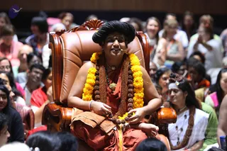 A seated figure in a large brown leather armchair with carved wooden arms is the central subject, wearing a tall black hair bun, a rust-orange robe, a long rudraksha mala and a thick yellow-orange marigold garland. A small clip microphone is on his face and his hands rest on the armrest. Behind him, a large audience fills the hall, mostly women and some men in pink, red, white and green clothing, many smiling or clapping. A woman on the right wears a white top, a long rudraksha-style necklace and a white headband with a small ornament. The hall has warm wood-panelled walls.