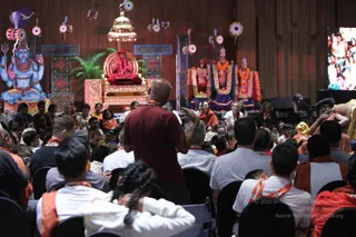 A photograph of a seated audience facing a stage in a wood-panelled hall. On the stage, a figure in red and gold sits on an ornate red throne beneath a gilded canopy, flanked by three deity murtis draped in garlands and colourful cloth. A blue painted backdrop shows a seated Shiva figure. In the foreground, a man with a shaved head in a maroon t-shirt stands with his back to the camera, gesturing toward the stage, while attendees in white and orange-saffron shawls and lanyards sit in rows of chairs. A large screen on the right shows images.