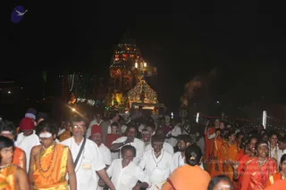 Night procession at a temple-style ratha, a decorated chariot lit with string lights and small lamps, pulled through a crowd. Many people walk in white dhotis and shirts, and others wear saffron and orange robes, with some in red and orange headwear. A man in the foreground wears an orange shawl and rudraksha beads, a woman in orange sari has a bindi, and a smoke plume rises near the chariot. Lit decorations line the route in the dark background.