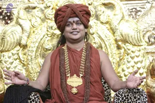 A man seated on an ornate gold throne with carved floral and scrollwork backrest, wearing a rust-orange turban wrapped in a braided crossover style, rust-orange robes draped over his shoulders, multiple rudraksha mala strands, and a heavy gold pendant necklace with a central gold medallion. He has a red bindi on his forehead, his hands are open with palms out in a gesturing pose, and he is speaking with a relaxed expression. The leopard-print fabric draped beside him is visible at the bottom. Swamiji (probable) based on the turban, saffron-orange robes, rudraksha malas and ornate gold regalia, seated in discourse.