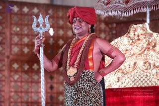 A standing man in a red turban and red shawl holds a tall silver trident in his right hand while his left hand rests on his hip. He wears layered rudraksha malas and a heavy gold ceremonial necklace with a pendant, bright red bangles on his arms, and a leopard-print lower garment. Behind him is an ornate gold throne with red velvet at its base, a carved wooden lattice wall, and a red-and-gold canopy at the top right.