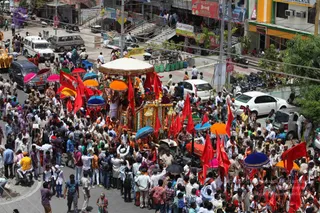 A street procession on a busy city road lined with shops, parked cars, motorbikes and pedestrians. At the centre, a decorated float with a white and gold canopy and a garlanded platform carries a figure in white seated near the front, surrounded by marigold garlands. Many red flags are held aloft, alongside orange, yellow and pink umbrellas. Uniformed police officers and men in white kurta-pyjama walk around the float, with crowds of spectators and devotees in saffron and white on both sides. Hanging signs and shop fronts show Hindi and English lettering.