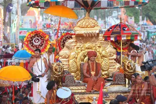 Ceremonial procession at a crowded street festival. Seated centrally on an ornate gilded palanquin throne with carved lion heads and leopard-print cushions is a man in saffron-orange robes, a saffron turban, and layered rudraksha and gold necklaces, hands resting in his lap. Behind him a bare-chested attendant in white dhoti and mala holds a fan and steadies the throne. Other attendants in saffron and white, some with shaved heads and malas, stand around the platform. A drummer in orange sits at the front with a large drum. Above and around are large umbrellas in orange, red and multicoloured fringed designs, with a crowd of onlookers and tents behind. A cameraman in white stands at left. The figure's identity cannot be confirmed from appearance, so no identification is made.