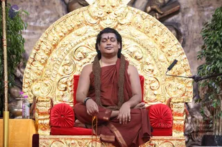 A seated man in a rust-red robe sits cross-legged on an ornate golden throne with red cushioned armrests and a gold arched back. His eyes are closed in meditation, with a red tilak on his forehead, a rudraksha-style mala of brown beads draped over his shoulders, and a red prayer mala in his hands. Potted green plants flank the throne, a microphone stand sits to the right, and a carved stone backdrop rises behind him. Pose and dress suggest a seated meditation or satsang, with a person candidate matching the saffron-red robe, mala and presence on the throne.