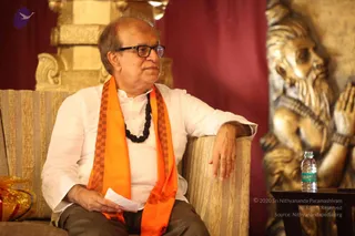 A seated man in a white kurta wears an orange and maroon checked shawl and a dark rudraksha-style bead mala, with glasses and grey trousers. He sits on a striped upholstered sofa with one arm resting on its armrest, holding a folded paper. Beside him stands a golden sculpture of a bearded sage with hands in anjali. A water bottle sits on a dark side table, and a gift wrapped in gold foil lies at left. Red drapes and a carved golden column form the backdrop.