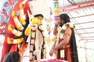 A standing man with long dark hair and a beard, bare-chested, wearing a rust-orange dhoti-style lower garment, a red and white flower garland and a gold armband, lifts his hand toward the face of a multi-armed deity murti. The murti has yellow skin, red and gold clothing, a white ornate crown and a trident-like staff held in one hand, with white floral decorations and red flower garlands. In the foreground, a metal lamp and a basin of pink flowers sit on the ground beside a partially visible person. The setting is a covered open pavilion with red painted pillars and a corrugated metal roof, with a colourful painted panel behind the murti.