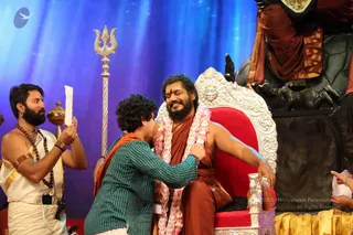 A photograph on a stage with a blue backdrop. A bearded man in an orange-brown robe with a pink-and-white flower garland sits smiling with eyes closed on an ornate silver and red throne decorated with a sun motif. A woman with curly hair in a green striped shirt and orange shawl leans in to embrace him and hold his arm. On the left, a man in a white dhoti and shawl, wearing a rudraksha mala and bangles, holds up a paper. A silver trident stands beside the throne and a dark carved backdrop with a deity figure appears at right.