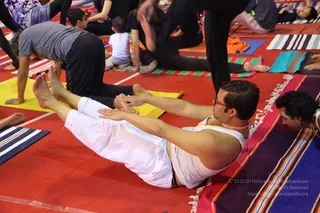 A yoga class held on a red floor covered with striped and coloured mats. In the foreground a man in a white sleeveless top and white trousers with a thin neck cord lies on his back, legs extended and arms reaching forward toward his feet in a seated forward-stretch posture. To his right a bearded man with curly hair lies on a striped mat, watching. Behind them, several participants in black clothing bend forward or sit with legs stretched on yellow, green and orange mats, and a small child sits on a mat at the back. No identifiable a person candidate is visible; the people are practitioners in asana poses.