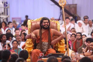 A seated man on an ornate gold-and-brown leather throne with golden lion armrests smiles broadly in front of a large seated audience. He wears an orange-saffron cloth, a leopard-print waist wrap, and a long rudraksha mala draped to his lap, with a small microphone at his cheek. Beside him stands a tall gold trident. Behind him, devotees in white and cream clothes, several in head wraps, sit under white tent walls with a sound mixer and cables visible. Swamiji (probable): long swept-back hair, ornate gold throne, saffron clothing and a rudraksha mala.
