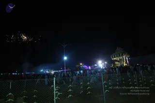 A night-time outdoor photograph of a large crowd gathered along a roadside behind a wire-mesh fence decorated with green leaf garlands. On the right, a decorated ornamental temple chariot or palanquin with a striped canopy and lit lamps stands among the people. Fireworks burst in the dark sky on the upper left, and street lights, colourful lights and small parasols line the route. Most people are seen from behind or at a distance, some in white clothing. The scene suggests a procession at night.
