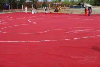 A photograph of a large red fabric ground with a white chalk or powder outline forming the sacred syllable Om, laid out in an open outdoor field. At the far end stands an ornate golden palanquin-style structure with a canopy and a golden deity murti on a red platform, flanked by brass lamp stands. Along the top edge, several people in white dhoti and shawl stand near a camera on a tripod, and a few people in white and one in orange clothing stand near the formation. On the right, a woman in an orange sari and another in a purple sari stand at the edge of the red field. Trees and a red boundary wall form the background.