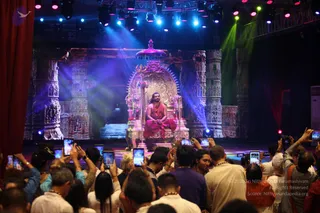 A stage event photograph with blue, purple and green stage lights. A seated man in a red-orange robe with a rudraksha mala sits on an ornate silver throne with a canopy and a trident beside him, before a backdrop of carved stone pillars. In the foreground, a crowd of devotees in white and orange clothing holds up smartphones to record him. The swami-like figure is seated centrally as the focus of the scene. Some attendees wear white garlands and orange shawls.