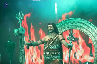 A man stands on a stage in front of a backdrop of orange and red flames lit in teal light. He wears a leopard-print garment with rows of brass bells across his chest and waist, rudraksha bracelets on his arms, a bindi on his forehead and a thin chain at his neck. Long dark hair falls past his shoulders. He holds a trident in his right hand and a damaru-shaped drum with a leather cord in his left, while a large ornate silver-green circular arch stands behind him. A tall ceremonial trident and a peacock-feather fan stand to the left.