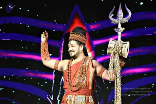 A performer stands on a stage against a dark backdrop with blue and pink light streaks and small sparkles. He wears a rust-orange wrap with a leopard-print sash, a crown-like headpiece of rudraksha beads over long dark hair, layered rudraksha malas, and beaded orange armbands. He raises his left arm and holds a silver trident wrapped with a leopard-print cloth in his right hand. A figure resembling a Shiva depiction is portrayed in this costume. a person: the long swept-back hair, rudraksha adornments and saffron-orange clothing match the appearance cues, though identity is uncertain from appearance alone. A red triangular light glows behind him.
