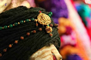 Close-up photograph of a person's head from the side, wrapped in a dark green turban of tightly wound fabric. Tied into the turban is an ornate gold ceremonial brooch with a carved relief figurative design and a red stone at its top, with green beads and gold spacer beads strung across the wrap. Dangling rudraksha seeds hang from the turban and gold clasp. The face in the lower corner is pale with a red marking near the cheek. The background is a soft blur of purple, orange and pink fabrics.
