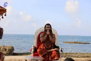 A man seated on an ornate silver-and-red throne on a sandy beach with the sea behind him, hands pressed together in namaskar. He wears saffron-orange robes, a red tilak on his forehead, long dark hair, and multiple strands of rudraksha mala beads around his neck and arms. A small silver side table stands in front of the throne. To the left, a partially visible attendant with white body markings and a red-and-gold canopy fringe. Swamiji (probable): the seated figure's long hair, saffron clothing and rudraksha malas match the cues for the description's appearance-based label.