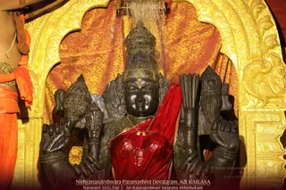 A dark stone deity murti with four arms stands in a gilded arched shrine against a gold and orange brocade backdrop. Water streams down over the statue's head during an abhishekam. It wears a red draped cloth and a gold necklace, and holds ritual objects in its hands. At the left edge, a partially visible person in orange garments and a beaded mala stands beside the shrine. The bottom shows the caption a person Devalayam, Adi KAILASA, Navaratri 2021, Day 2 - Sri Rajarajeshwari Snapana Abhishekam.