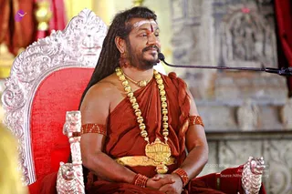 A man with long dark hair swept back and a tilak on his forehead sits on an ornate silver-and-red throne with carved lion-head arms, hands folded in his lap. He wears a rust-orange shawl draped over his bare shoulders, a heavy gold bead necklace with a large gold pendant, a gold waist belt, and red beaded armlets. A microphone on a stand is in front of him, and a carved stone wall stands behind the throne.