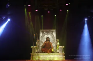A seated man in saffron-orange robes sits cross-legged on an ornate silver throne with lion figures flanking its back and armrests, on a gold-trimmed platform on a dark stage. He wears a rudraksha mala and a ceremonial gold necklace, with a red forehead mark, and raises his right hand in a blessing gesture. Two tall silver staffs stand beside the throne. Green, blue and magenta stage lights beam down from above.