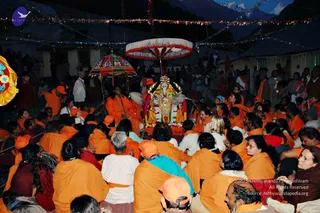 Night procession in a Himalayan town with many devotees seated on the ground in orange shawls and saffron clothing. At center, a decorated palanquin-style murti of a deity in gold and red garments and garlands is carried under two ornate umbrellas, one red-and-white and one multicolored. A man in orange robes stands near the murti, holding a fan-like chamara. Strings of lights hang across the scene and snow-capped mountains rise in the background. Many people in orange and white stand and sit around the procession.