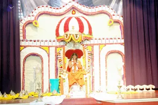 A seated figure in saffron-orange robes sits on an ornate throne at the centre of a stage decorated with white and red flower arrangements, scalloped arches and yellow marigold garlands. The seated person has hands pressed together in namaskar and a garland of white flowers draped over the shoulders. A red velvet backrest sits behind the throne, with a silver umbrella-shaped canopy above. Silver kalasha-style vessels and small brass oil lamps lie on the floor around the base, and a teal bucket and a standing fan appear at the sides, with purple curtains framing the stage. The setting suggests a guru darshan or satsang during a Guru Poornima event.