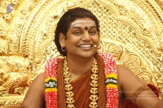 Close photo of a smiling man seated before an ornate gold repoussé backdrop. He wears a white forehead mark, a red bindi, and gold hoop earrings, with a small clip-on microphone near his mouth. He has a multi-coloured garland of pink, yellow and white flowers over an orange-brown cloth, layered with gold chain necklaces and rudraksha-style beads, plus a gold armlet. Swamiji (probable): long swept-back hair, ornate gold jewellery and orange-saffron dress with a rudraksha mala.