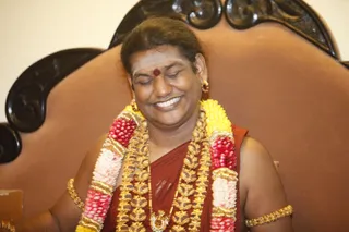 A smiling man with closed eyes sits on a brown upholstered throne with carved dark wooden arm panels. He wears a red-maroon garment over bare shoulders, heavy gold ornate necklaces, gold armlets and bangles, and a garland of yellow and pink-red flowers draped over his shoulders. A red dot marks his forehead, with white markings beneath it. Only one person is visible.