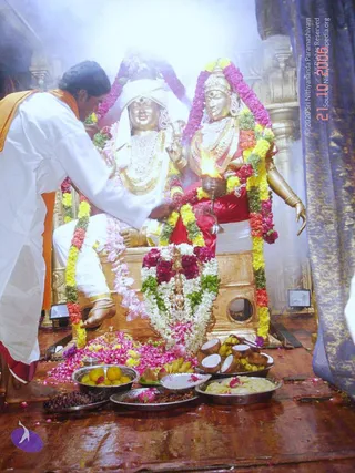 A photograph of a ritual worship scene before two golden deity murti seated on a gilded throne, decorated with garlands of yellow, red, pink and white flowers. The murti on the left wears a white cloth and white jewelled necklaces, and the one on the right wears a red garment and an ornate gold necklace while holding a lit oil lamp. A man in a white kurta with an orange shawl stands at left, bending to perform padapuja by touching the feet of the seated white-clad murti. Smoke fills the air. In front lie plates of coconut halves, bananas, citrus fruits, sweets, pulses and rose petals, with a scattering of flower petals on the wooden floor.