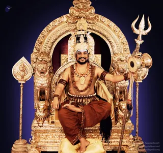 A seated figure on an ornate gilded throne with carved lion supports and an arched gold canopy against a deep blue backdrop. The person is bare-chested, wearing a rust-orange dhoti with a tiger-striped cloth, a wide dark beaded waist belt, multiple rudraksha malas across the chest and arms, and a large ceremonial pendant necklace. A tall gold crown and blue forehead markings are visible. He holds a silver trident in his right hand and a damaru drum with a long tassel. Gilded lion figures flank the seat, and a golden kalasha-style finial rises on the left. Appearance-based candidate a person seated on a peetha throne, presented as the central figure, with the throne and regalia prominent.