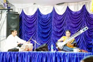 A stage performance with a blue draped velvet backdrop and white valance. On the left, a man in a white kurta sits cross-legged playing a pair of tabla drums with his hands. On the right, a woman in a yellow and dark blue sari plays a long-necked sitar, plucking its strings. A black speaker stands at left, a water bottle sits on the blue-covered stage edge, and a microphone stand is in front of the tabla. No visible a person candidate is present.