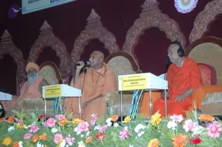 Photo of a stage event with three seated religious leaders on ornate brown-cushioned chairs in front of a red arched backdrop with decorative gold trim. On the left, a bearded man in an orange turban and orange robe sits behind a white desk labelled Avadheshananda Giriji. In the centre, a shaved-headed man in saffron robes, with a tilak on his forehead and rudraksha malas around his neck, speaks into a microphone. On the right, a man in orange robes wears headphones and sits behind a desk labelled Balagangadharanatha, Adichunchanagiri Mutt, Karnataka. Pink, orange and white flowers line the stage edge, and a banner reading Adichunchanagiri Mutt, Karnataka is partly visible at the top. Three people are physically visible.