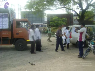 A photograph of a dusty roadside lot on a flood relief day. An orange Eicher truck on the left has a white banner with Kannada text on its side and a driver visible in the cab. Several men in white shirts and dark trousers stand near the truck, and a man in a checked shirt and dark trousers stands in the centre. A woman in a white salwar kameez with a white dupatta stands beside a silver minivan, and a man in a white and pink shirt opens the van's rear door. A man in a green shirt and khaki trousers holds a camera in the background, and a motorbike is parked at the right edge. Bare trees and a billboard appear behind them.