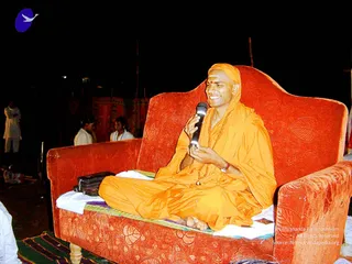 A seated man in saffron-orange robes and a matching saffron head wrap sits cross-legged on a red velvet sofa with leaf-patterned upholstery at night. He wears a red tilak on his forehead, a rudraksha mala around his neck, and a wristwatch, and smiles while holding a black handheld microphone in both hands. A dark leather bag rests beside him on a white cloth over the sofa. Two figures in white clothing stand in the background near a red draped backdrop and wooden poles. A woven striped mat lies on the ground in the foreground.