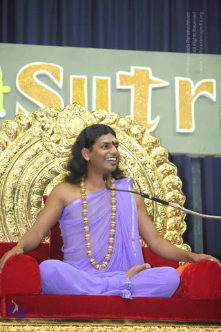 A seated man in a lavender-purple wrap and matching dhoti sits cross-legged on a red velvet throne with ornate gold carved back and armrests, speaking into a microphone on a flexible stand. He wears a long necklace of gold beads and a red bindi, and his long dark hair falls to his shoulders. Behind him a grey banner with large gold glittering lettering reading Sutra is displayed on a navy blue curtain. A second figure is not visible. This appears to be a talk or satsang setting, with the seated speaker as the central subject.