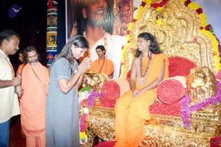 A photograph of a devotee in a grey kurta with hands joined in namaste, bowing before a seated figure on an ornate gold throne with lion-head armrests and red cushions decorated with marigold and purple garlands. The seated figure wears saffron-orange robes, a rudraksha mala and a long ceremonial pendant necklace, and has long dark hair. Behind them a large banner portrait shows a smiling man with long hair. An older bearded man in saffron robes and another saffron-clad attendant stand nearby; a man in cream clothing stands at left. Colourful painted pillars and a teal-and-purple stage backdrop frame the scene.