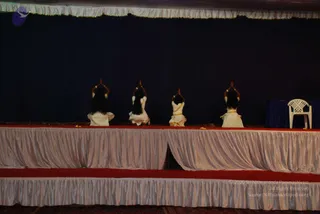 Photograph of a stage during a festival event. Four women with long dark hair, seen from behind, kneel in a row on a draped stage, arms raised with palms pressed together in namaste above their heads. They wear white and cream traditional dresses with gold-coloured borders. A white plastic chair sits at the right edge beside a dark blue draped table, with scattered flower petals on the red-carpeted stage. The backdrop is dark navy fabric under a pale draped canopy.